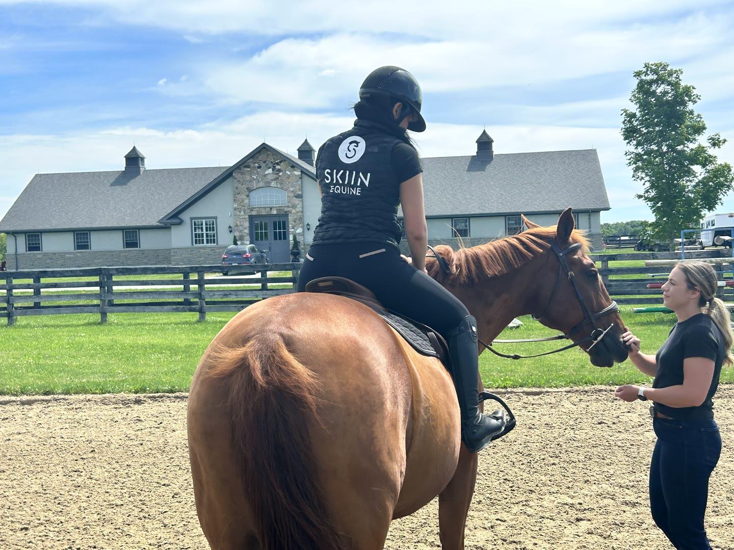 A rider wearing a SKIIN Equine vest mounted on a chestnut horse in an arena, a handler standing at the horse’s head